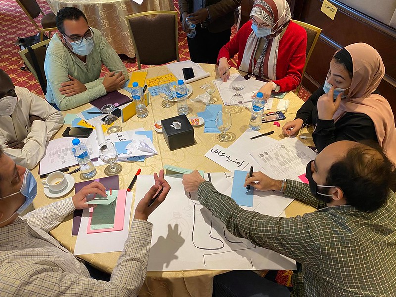 A group people sitting around a table having a discussion with pen, notes and coloured paper card. One of them is showing some card with writing to the others.